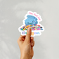 A hand holding a cute blue dragon sticker surrounded by colorful embroidery floss with the text "Floss Hoard Level Dragon"