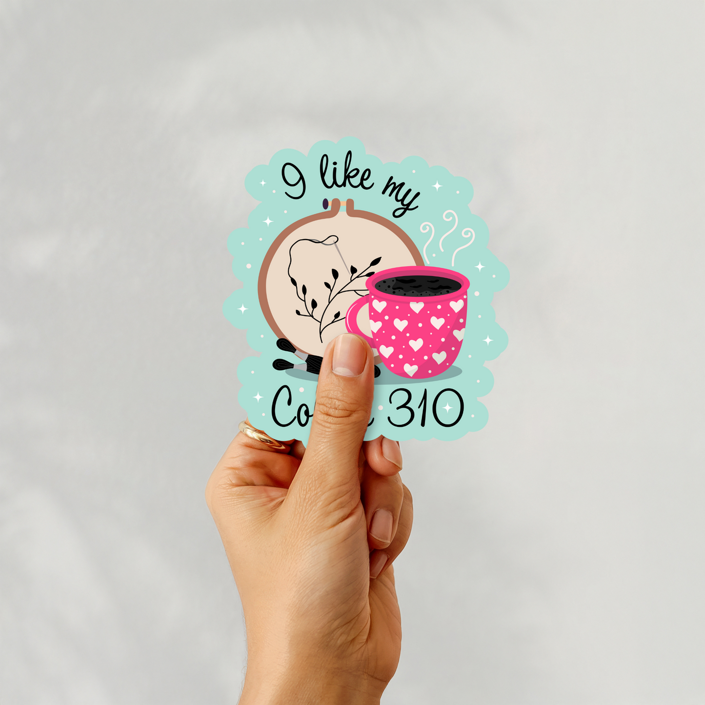 Hand holding ‘I Like My Coffee 310’ cross stitch design sticker with coffee mug and hoop art