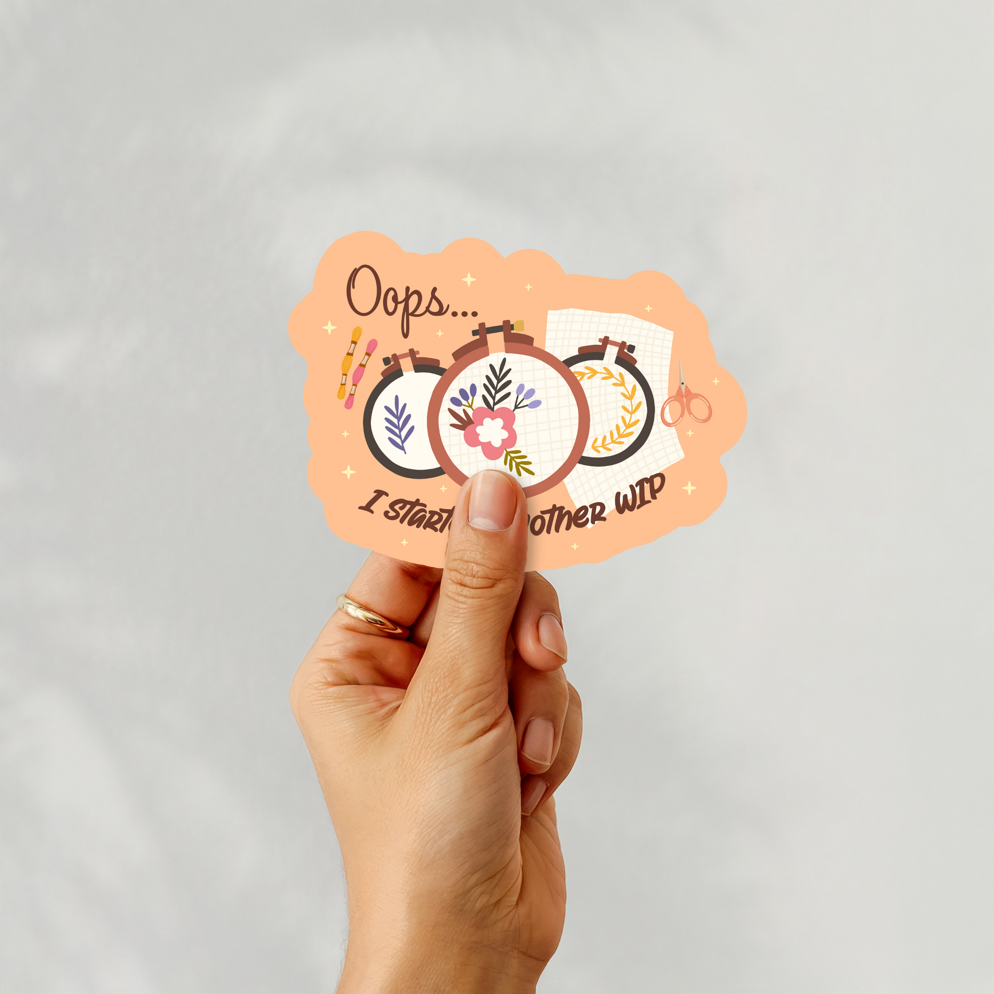 Hand holding ‘Oops... I Started Another WIP’ sticker featuring embroidery hoops and scissors